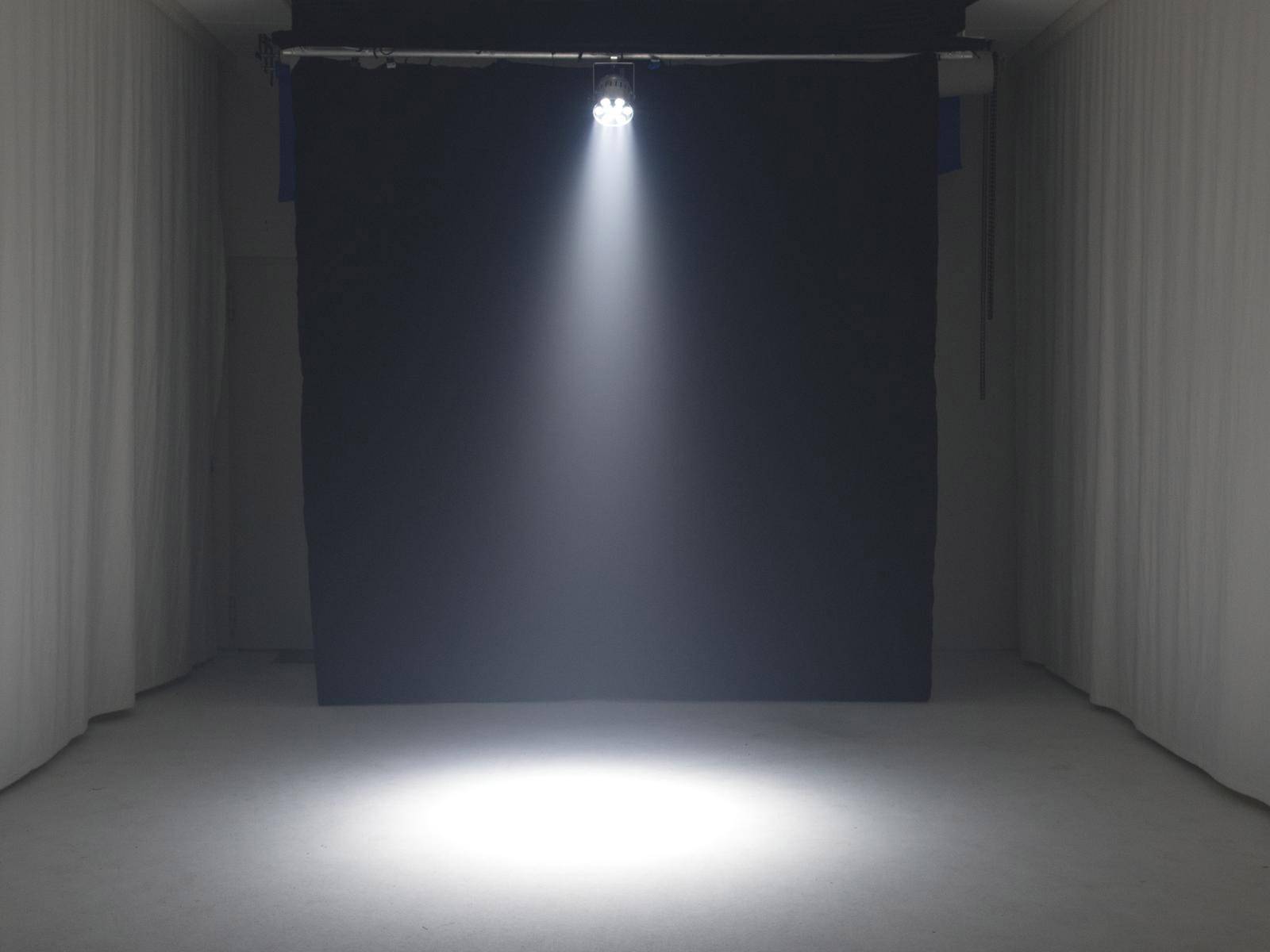 Samotny reflektor oświetla pustą, szarą scenę w ciemnym pomieszczeniu otoczonym białymi zasłonami.