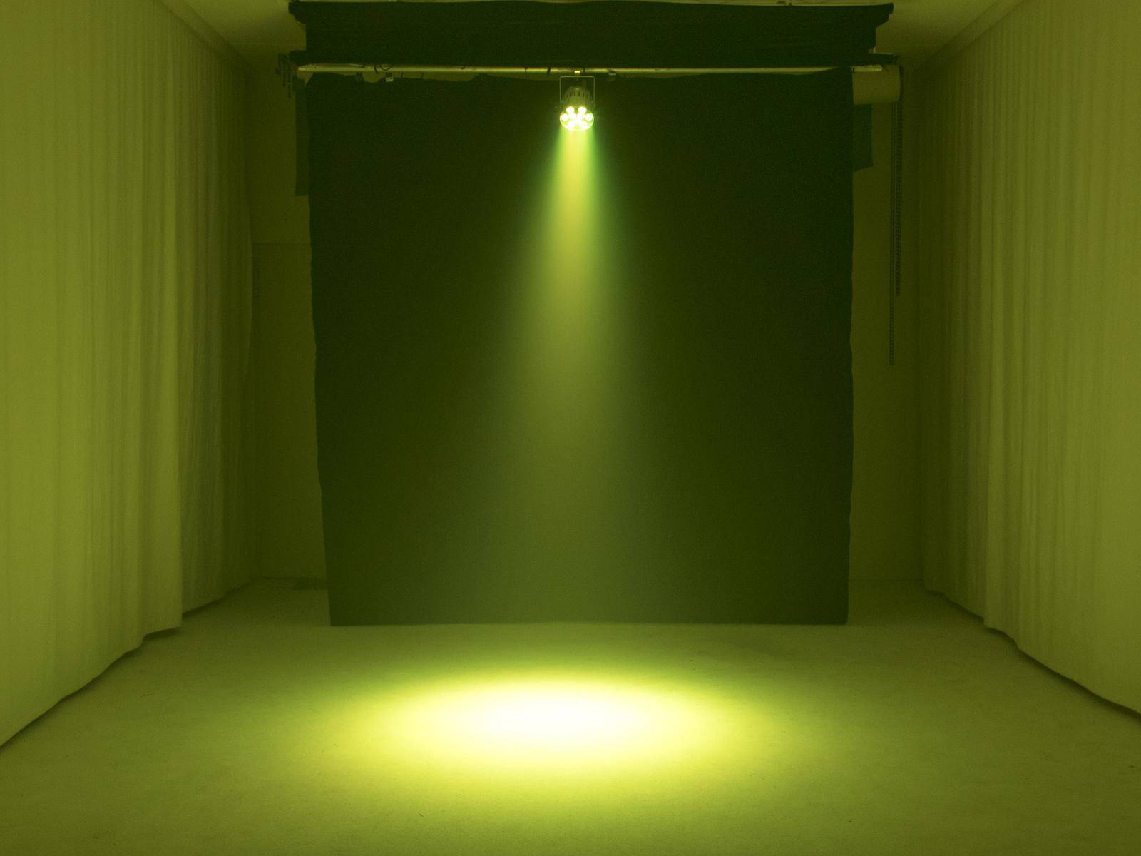 Jasny, żółty reflektor świeci z góry w ciemnym pomieszczeniu na pustą podłogę, tworząc okrągłą plamę światła.