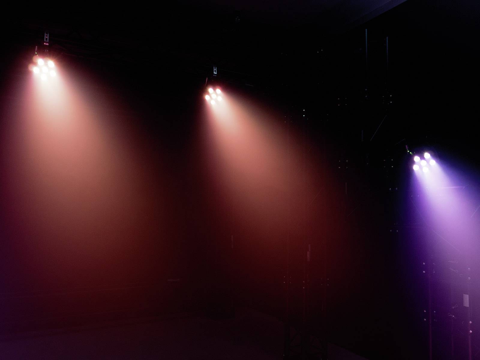 Trzy kolorowe reflektory oświetlają ciemną scenę. Po lewej stronie świecą na czerwono i fioletowo, po prawej - fioletowo. Brak widocznych osób.