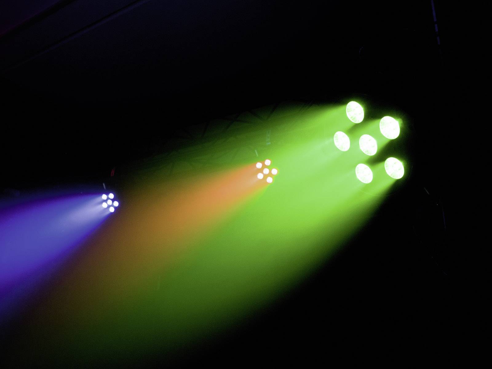 Kolorowe reflektory promieniują w odcieniach niebieskiego, pomarańczowego i zielonego, przesuwając się od prawej do lewej w ciemnym pomieszczeniu, tworząc żywe efekty świetlne.