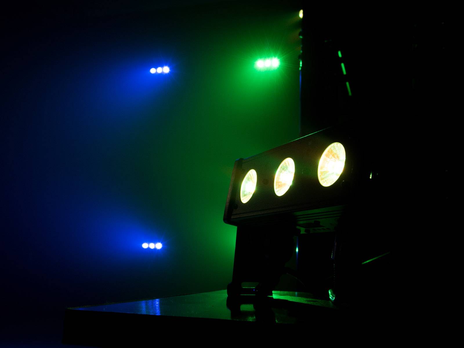 Wielokolorowe światła sceniczne w ciemnym otoczeniu, z przodu trzy reflektory, z tyłu sześć świateł w odcieniach niebieskiego i zielonego.