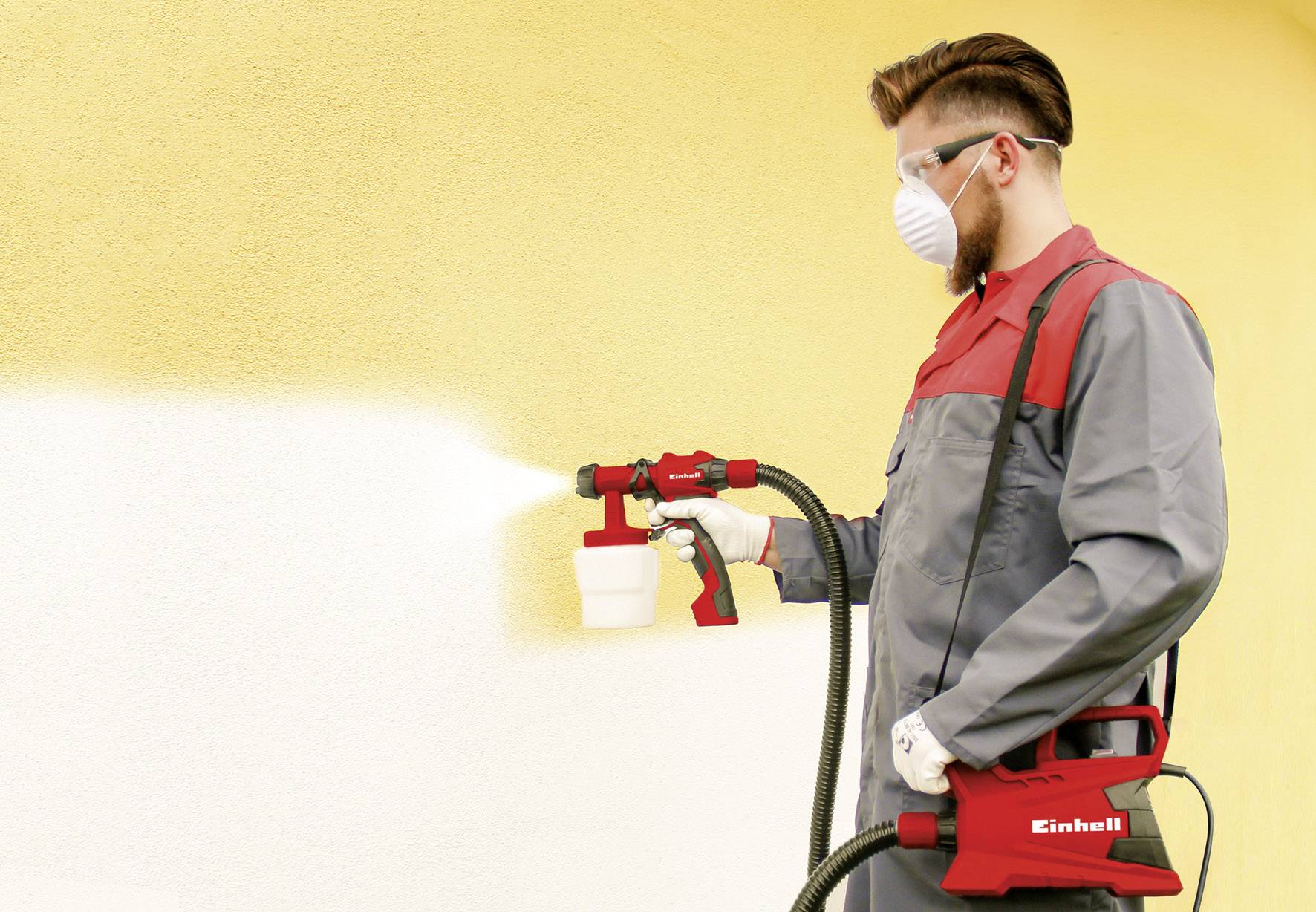 Mężczyzna rozpyla żółtą farbę na ścianę za pomocą pistoletu natryskowego. Jest ubrany w maskę ochronną, okulary ochronne i odzież roboczą.