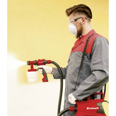 Mężczyzna w okularach ochronnych i masce nakłada żółtą farbę za pomocą systemu natryskowego na ścianę. Ma na sobie szare ubranie robocze.