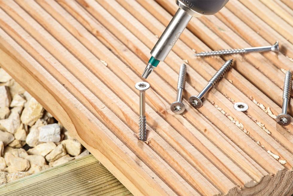 Zbliżenie wkrętarki akumulatorowej wkręcającej wkręt w deskę drewnianą na placu budowy. Obok leżą kolejne wkręty.