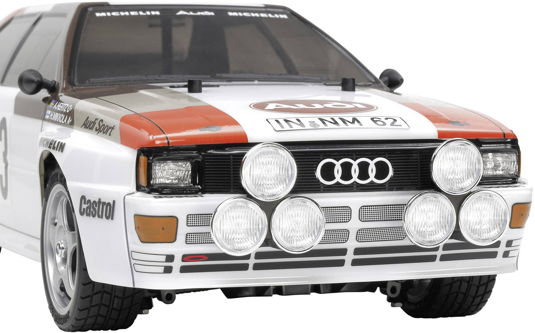Biały samochód rajdowy Audi Quattro z charakterystycznymi reflektorami i numerem rejestracyjnym 'IN-NM 62', ozdobiony logo sponsorów.