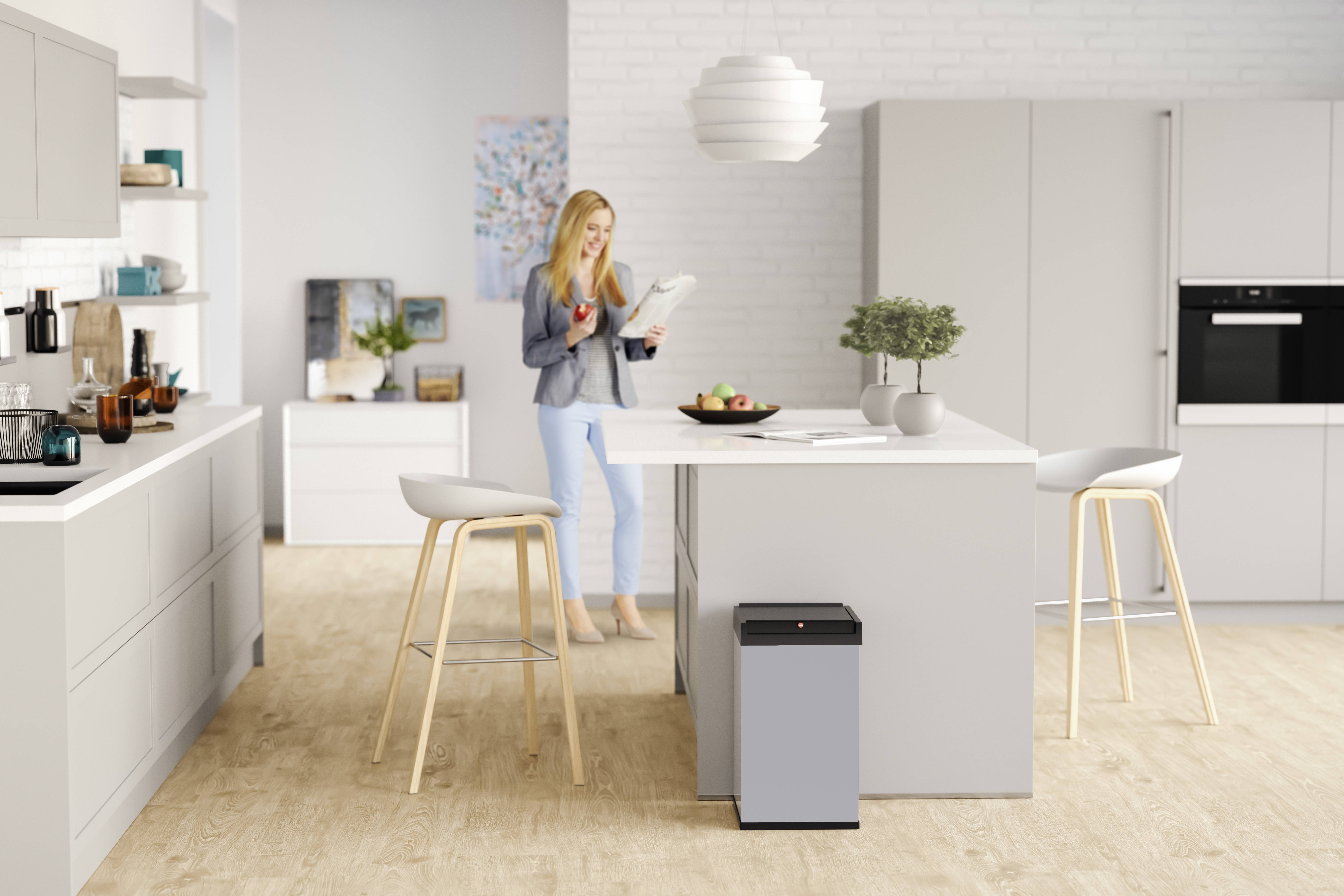 Kobieta stoi w nowoczesnej kuchni i czyta książkę. Trzyma czerwony kubek, w tle widoczna jest biała szafka i rośliny.