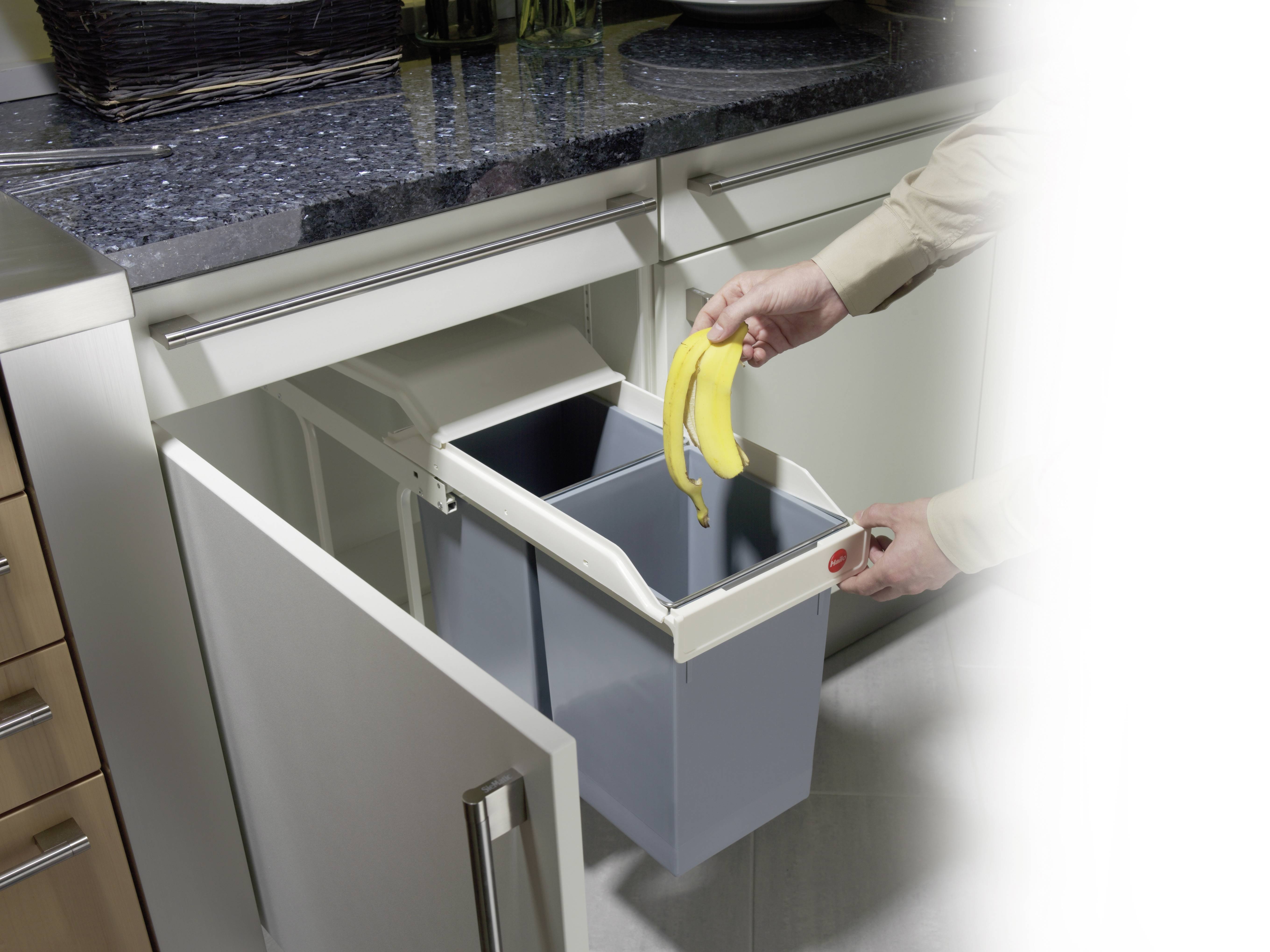 Osoba wrzuca skórkę banana do wysuwanego kosza na śmieci w kuchni pod blatem roboczym.