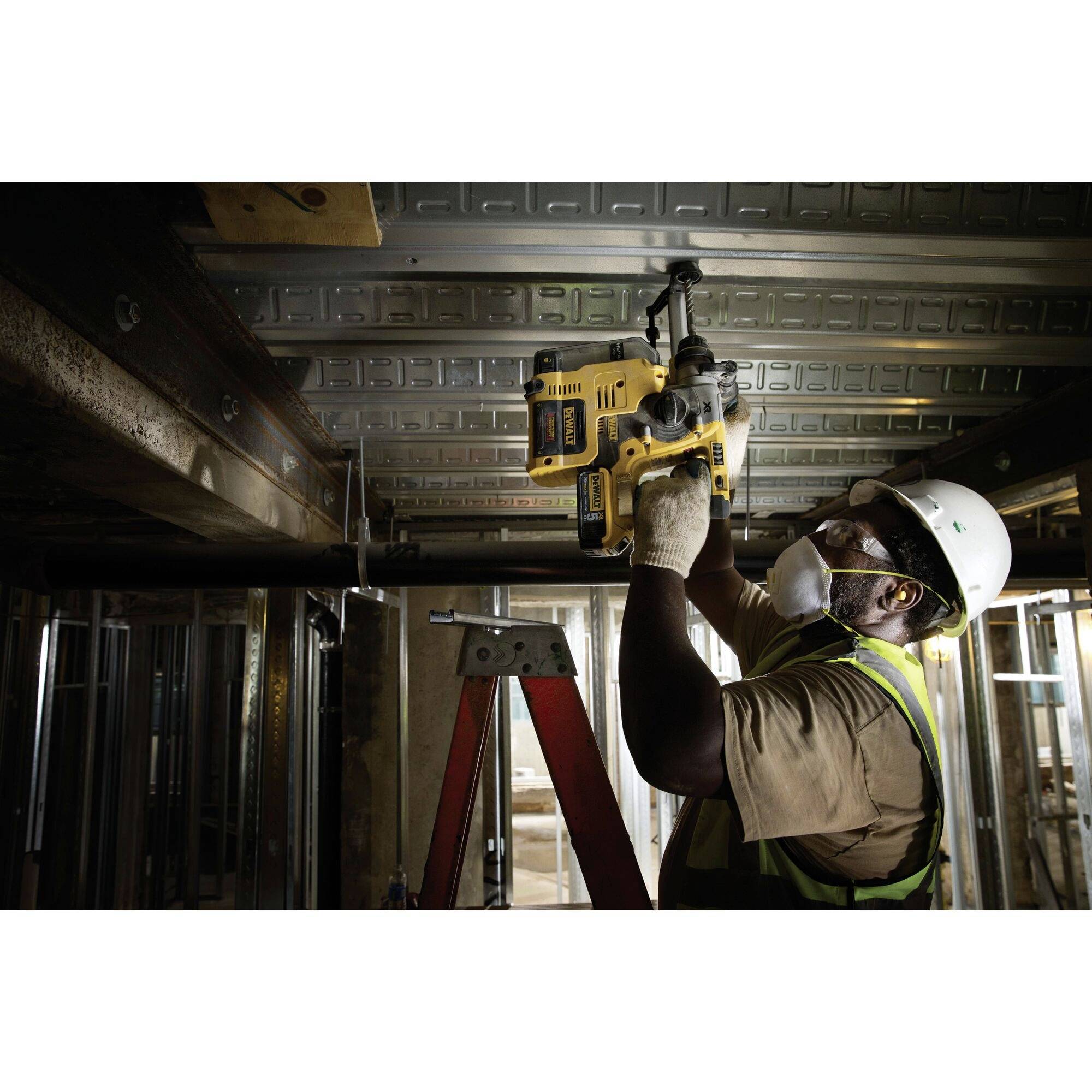 Robotnik budowlany w ubraniu ochronnym wierca otwór w suficie w niedokończonym budynku za pomocą elektronarzędzia.