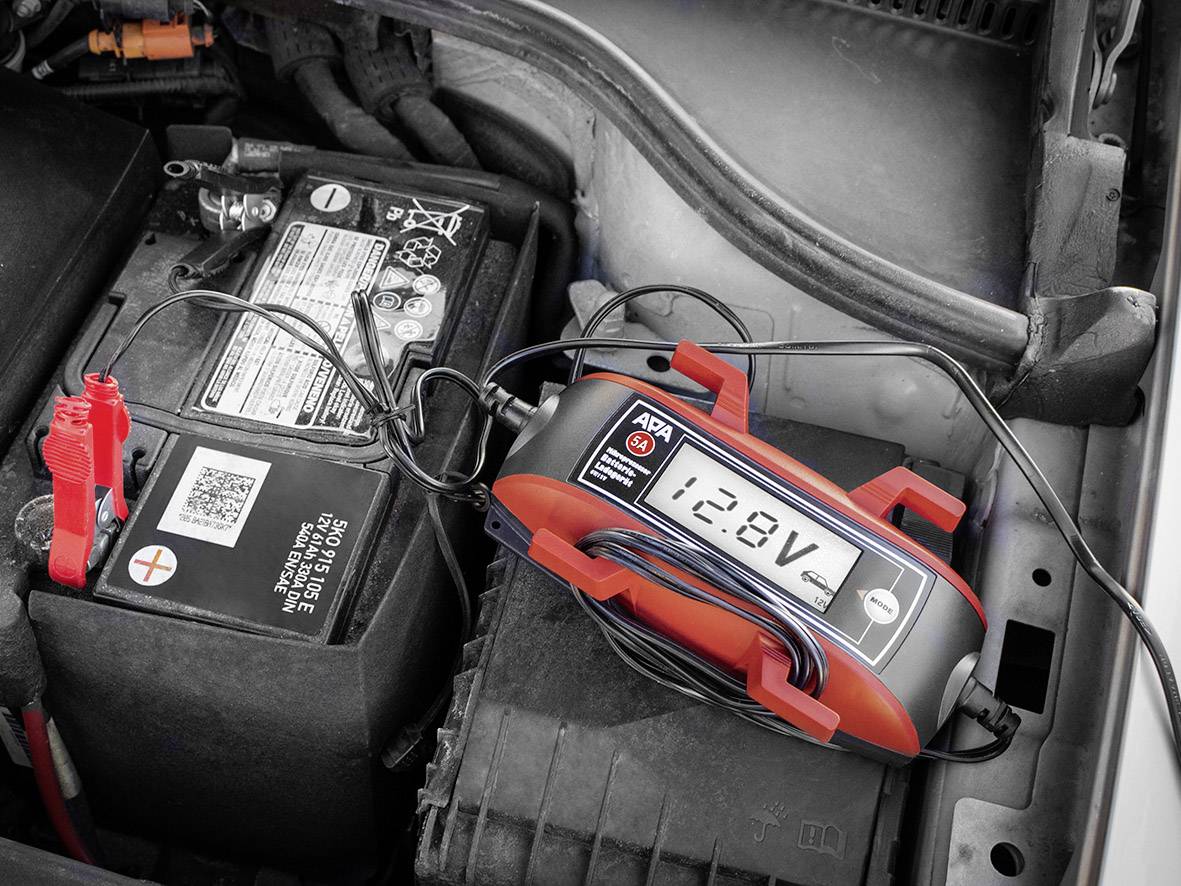 Ładowarka '12,8V' podłączona do akumulatora samochodowego w komorze silnika, wskazująca napięcie podczas ładowania.
