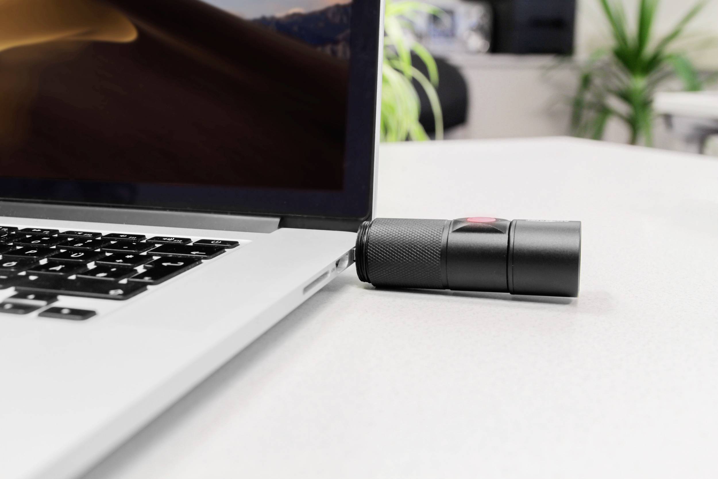 Laptop stoi na stole. Mały, czarny głośnik lub podobne urządzenie opiera się o prawą stronę laptopa.