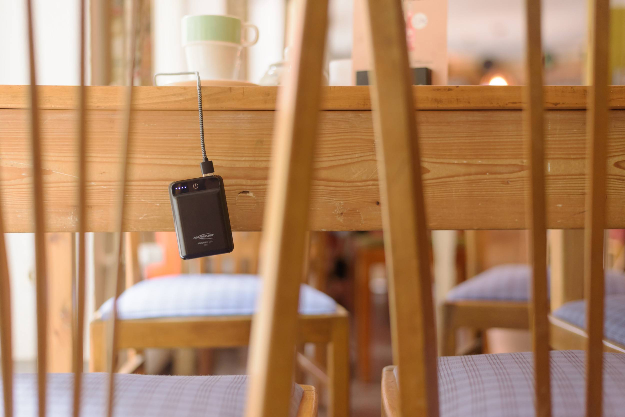 Smartfon zwisza odwrócony do góry nogami na przewodzie ładującym pod drewnianym stołem w przytulnym pomieszczeniu z krzesłami i kubkami.