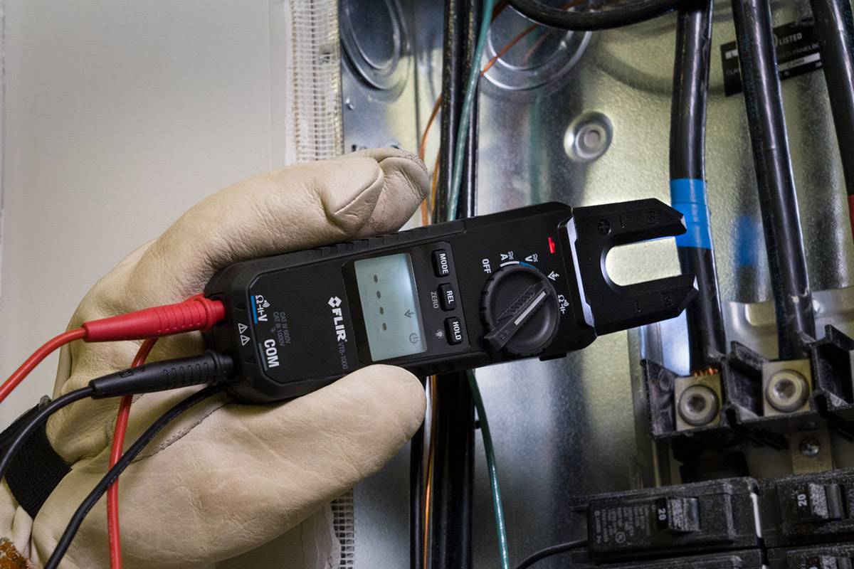 Dłoń w rękawicy trzyma elektryczny miernik nad przewodami w skrzynce bezpieczników. Wyświetlacz pokazuje dane pomiarowe.