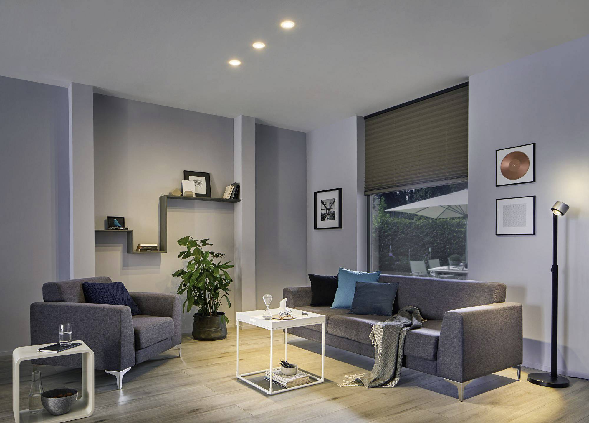 Nowocześnie urządzony salon z szarą sofą, fotelem, stolikiem pomocniczym i rośliną doniczkową. Miękkie oświetlenie tworzy przytulną atmosferę.