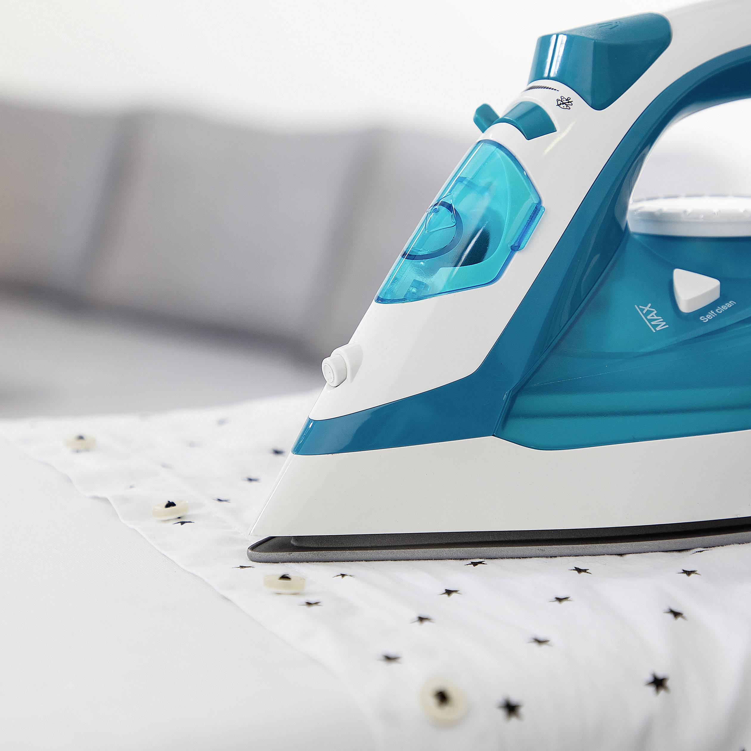 Żelazko prasuje białą koszulę na desce do prasowania. Koszula ma wzór małych gwiazdek. W tle niewyraźnie widać poduszki na kanapie.