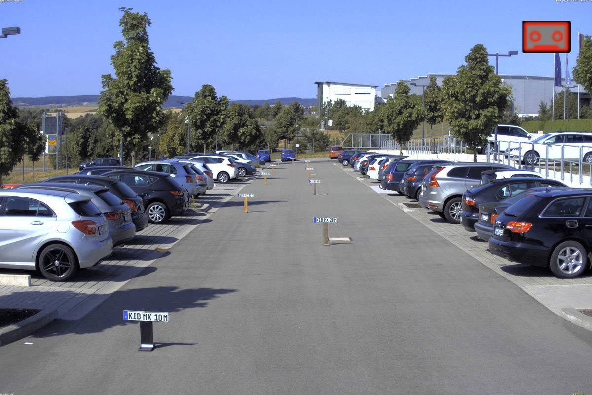 Parking z wieloma zaparkowanymi samochodami po obu stronach i zielonymi drzewami w tle.
