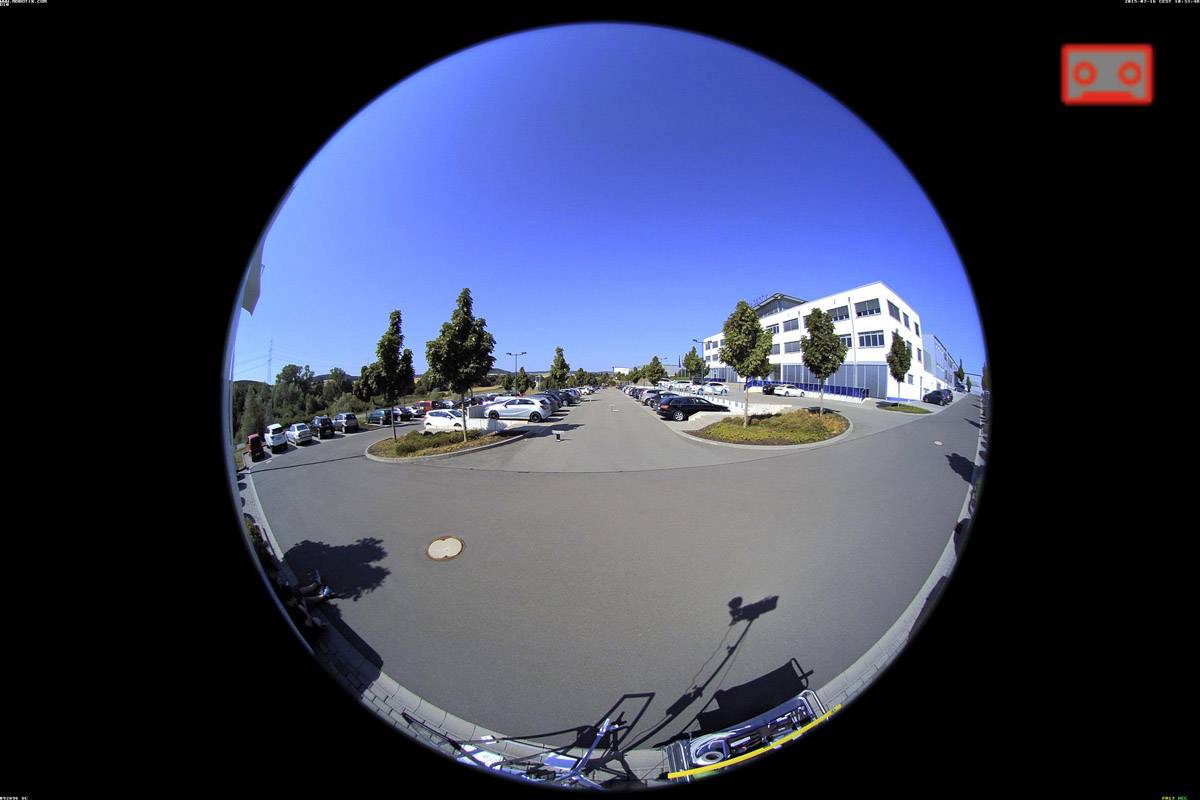 Okrągłe ujęcie ulicy z zaparkowanymi samochodami, otoczonej budynkami i drzewami na tle błękitnego nieba.