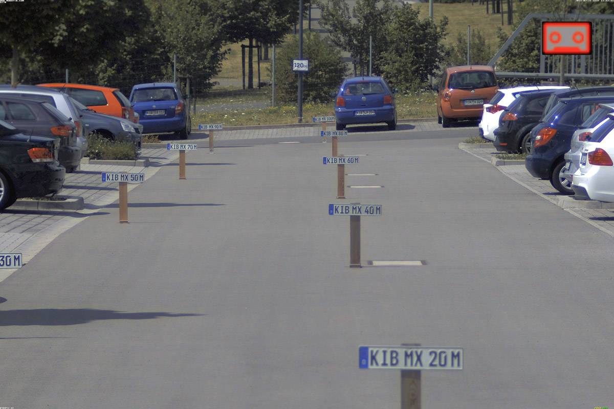 Kilka znaków drogowych na parkingu wskazuje odległość do 'K1B MX 20M'. Tablice są ustawione wzdłuż drogi.