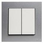 Podwójny przełącznik kołyskowy w srebrnej ramce zamontowany na ścianie, służący do sterowania urządzeniami elektrycznymi lub oświetleniem.