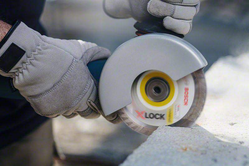 Osoba nosi rękawice robocze i używa szlifierki kątowej z tarczą 'X-LOCK' do cięcia powierzchni kamiennej.