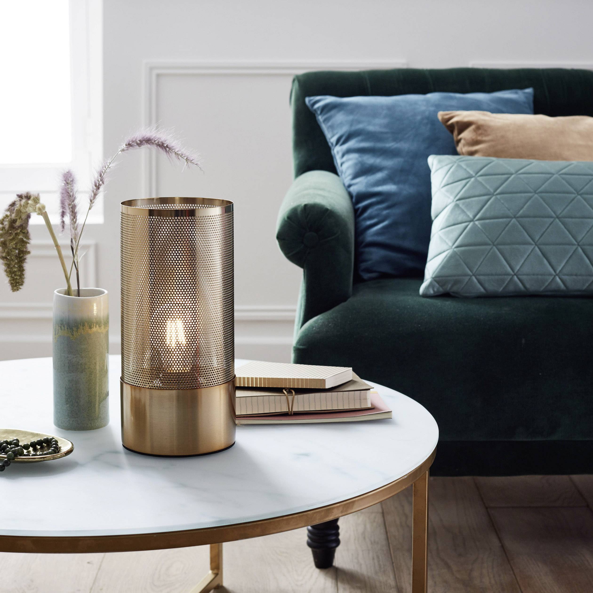 Stolik z lampą z metalowej siatki, obok wazon z trawami i stos książek, przed zieloną sofą ozdobioną kolorowymi poduszkami.