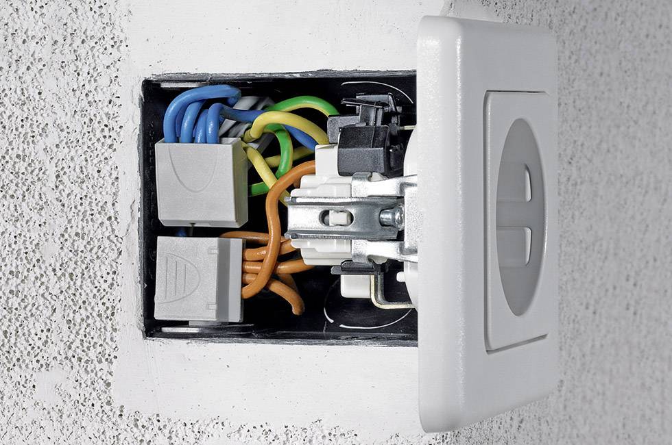 Otwarte gniazdo elektryczne w ścianie z widocznymi, kolorowymi przewodami i dwoma szarymi skrzynkami rozdzielczymi.