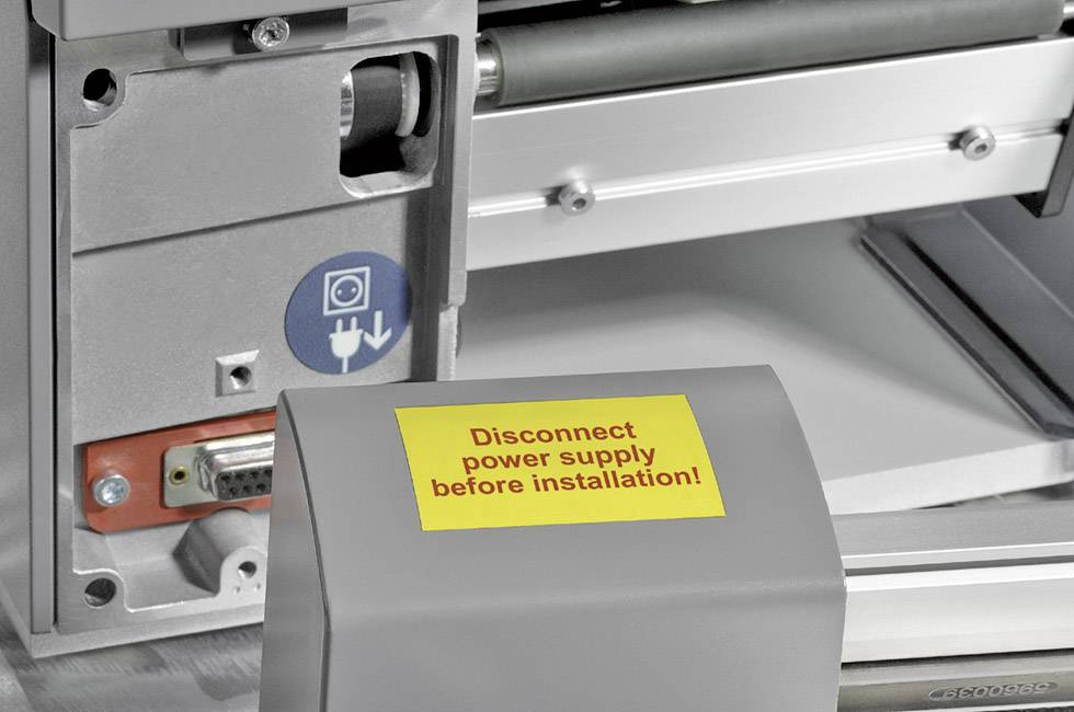 Na żółtej tabliczce widnieje napis: 'Przed montażem odłączyć zasilanie'. Obok znajdują się komponenty maszyny z symbolem ostrzeżenia bezpieczeństwa.