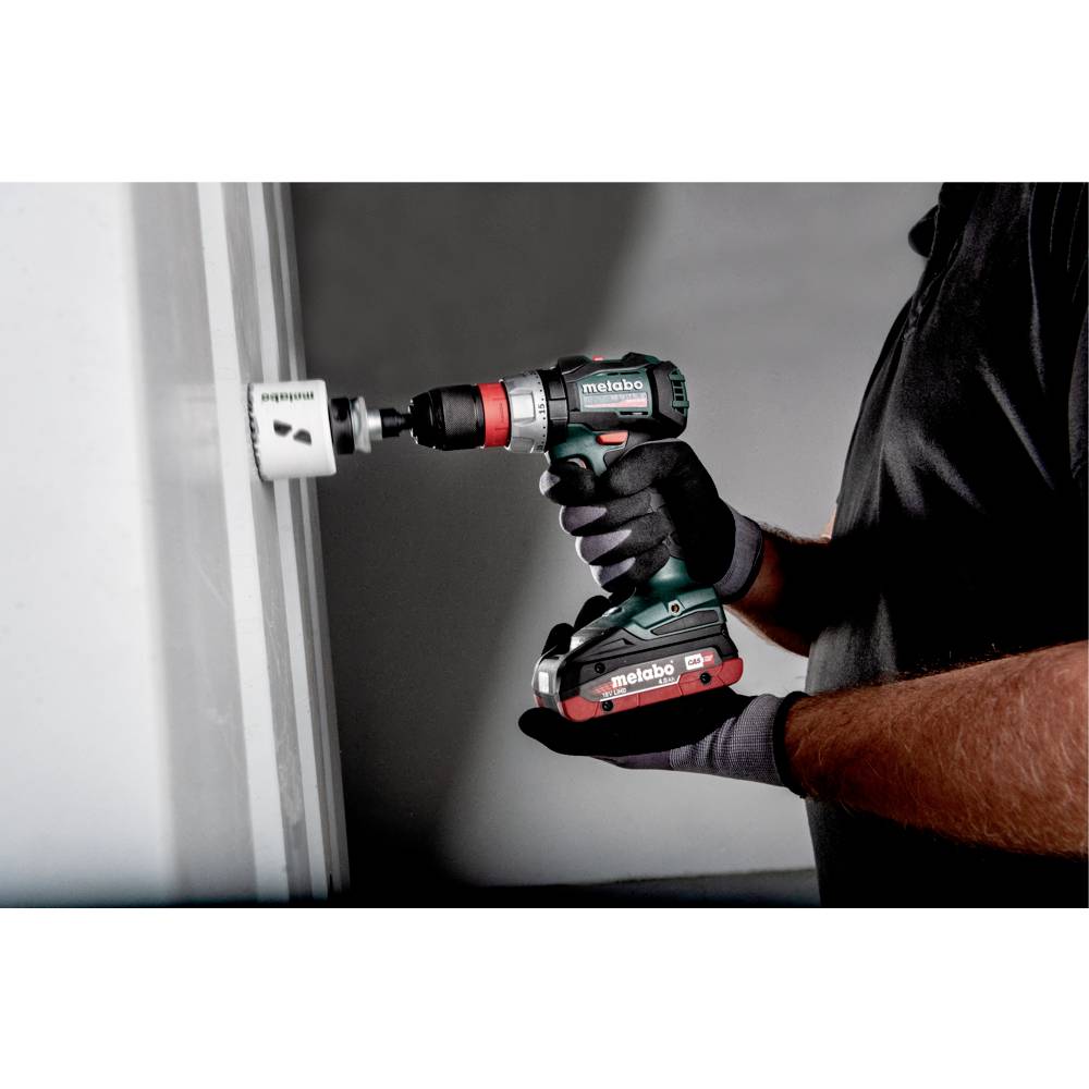 Osoba używa bezprzewodowej wiertarki, aby zrobić otwór w białej ścianie. Wiertarka jest czarna i zielona, z logo marki z boku.