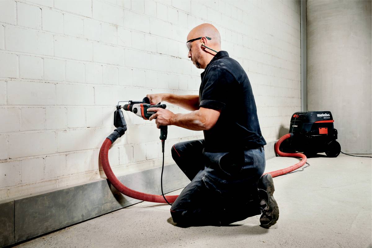 Mężczyzna klęczy na podłodze i wywierca otwór w ścianie z cegły, z odkurzaczem w tle, który usuwa pył.