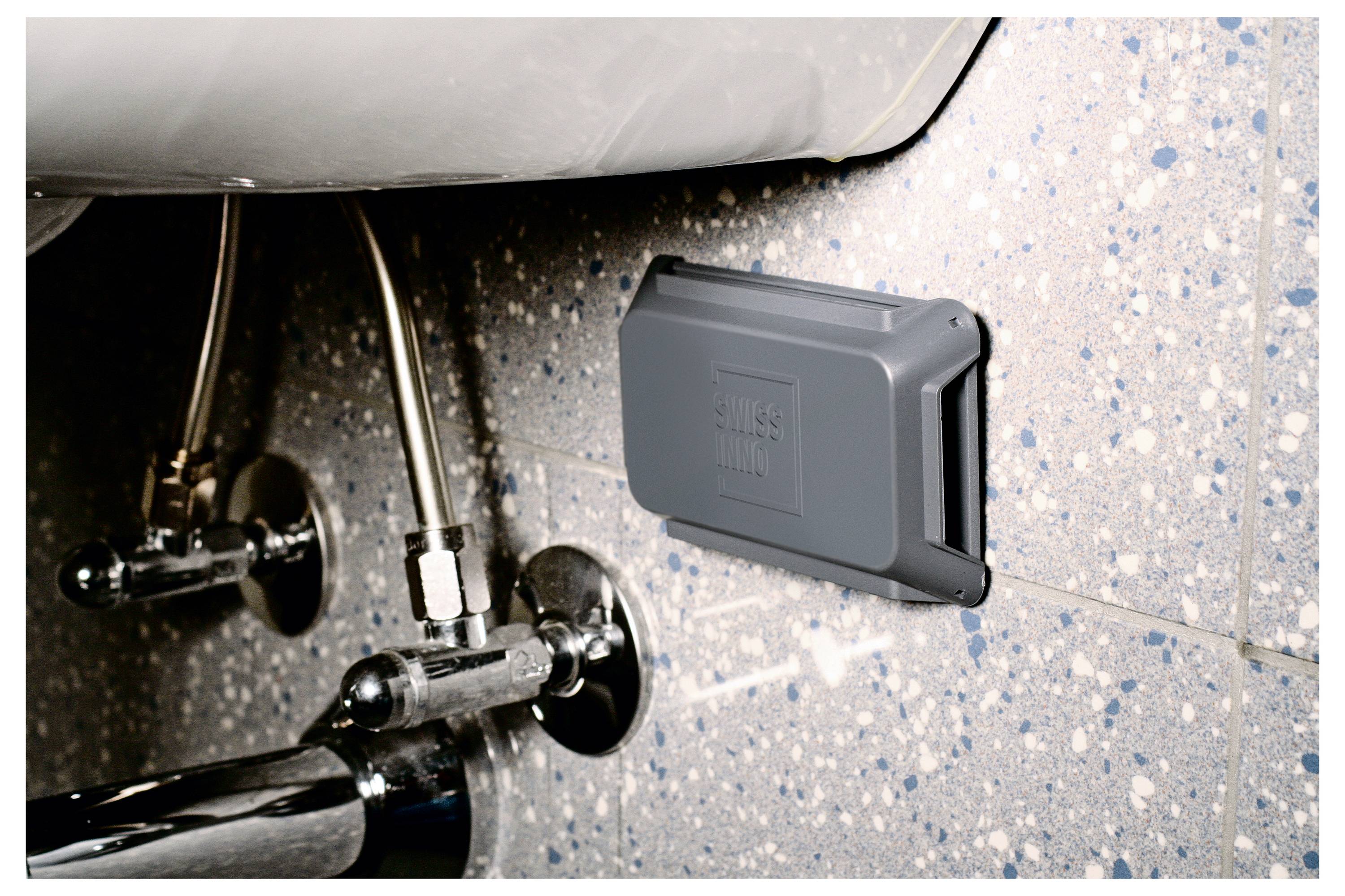 Szary plastikowy przyrząd zamontowany na nakrapianej ścianie łazienki, w pobliżu armatury sanitarnej pod zlewem.