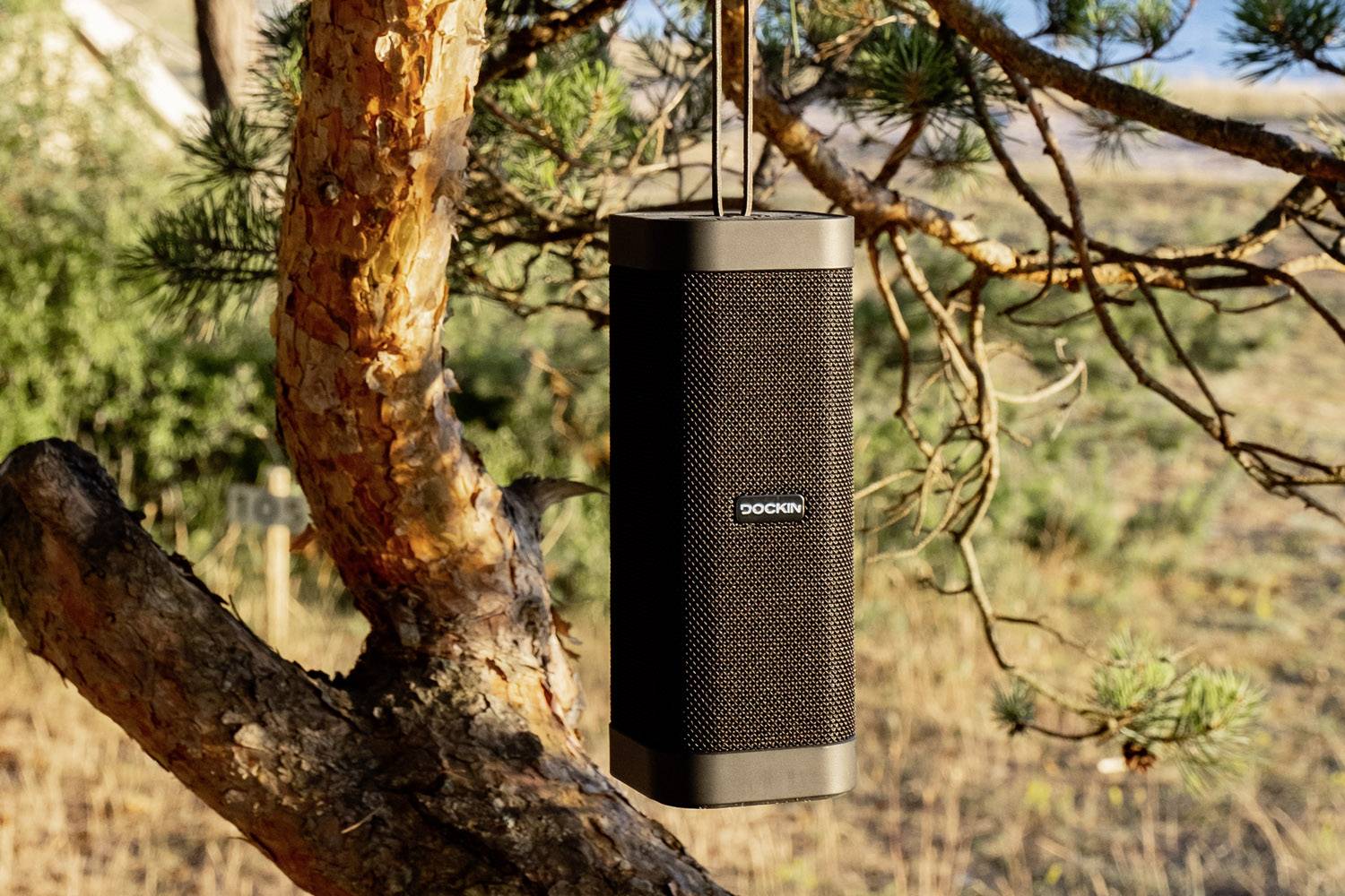 Czarny, przenośny głośnik wisi na drzewie na wolnym powietrzu, otoczony przyrodą i słonecznym światłem.