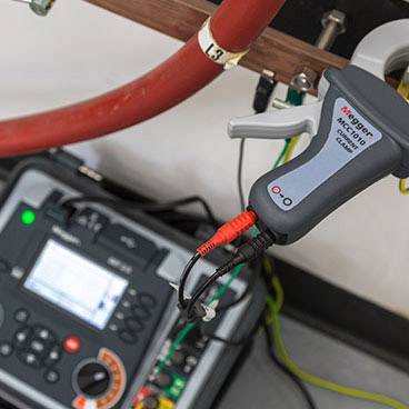 Multimetr i zaciski na czerwonym przewodzie, prawdopodobnie używane do pomiarów elektrycznych, z niewyraźnym przyrządem pomiarowym w tle.