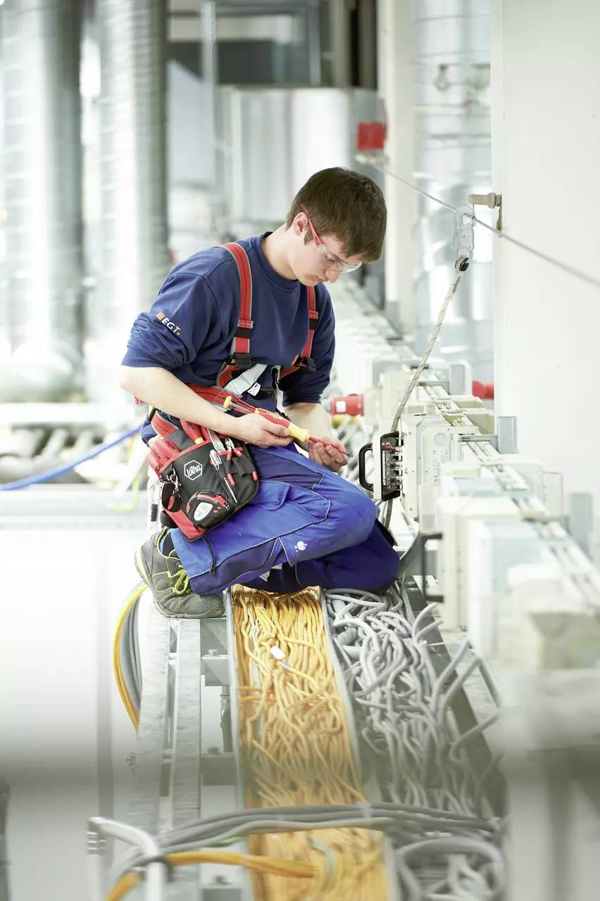 Technik w odzieży roboczej klęczy na kanale kablowym i pracuje przy rozdzielnicy elektrycznej w środowisku przemysłowym.