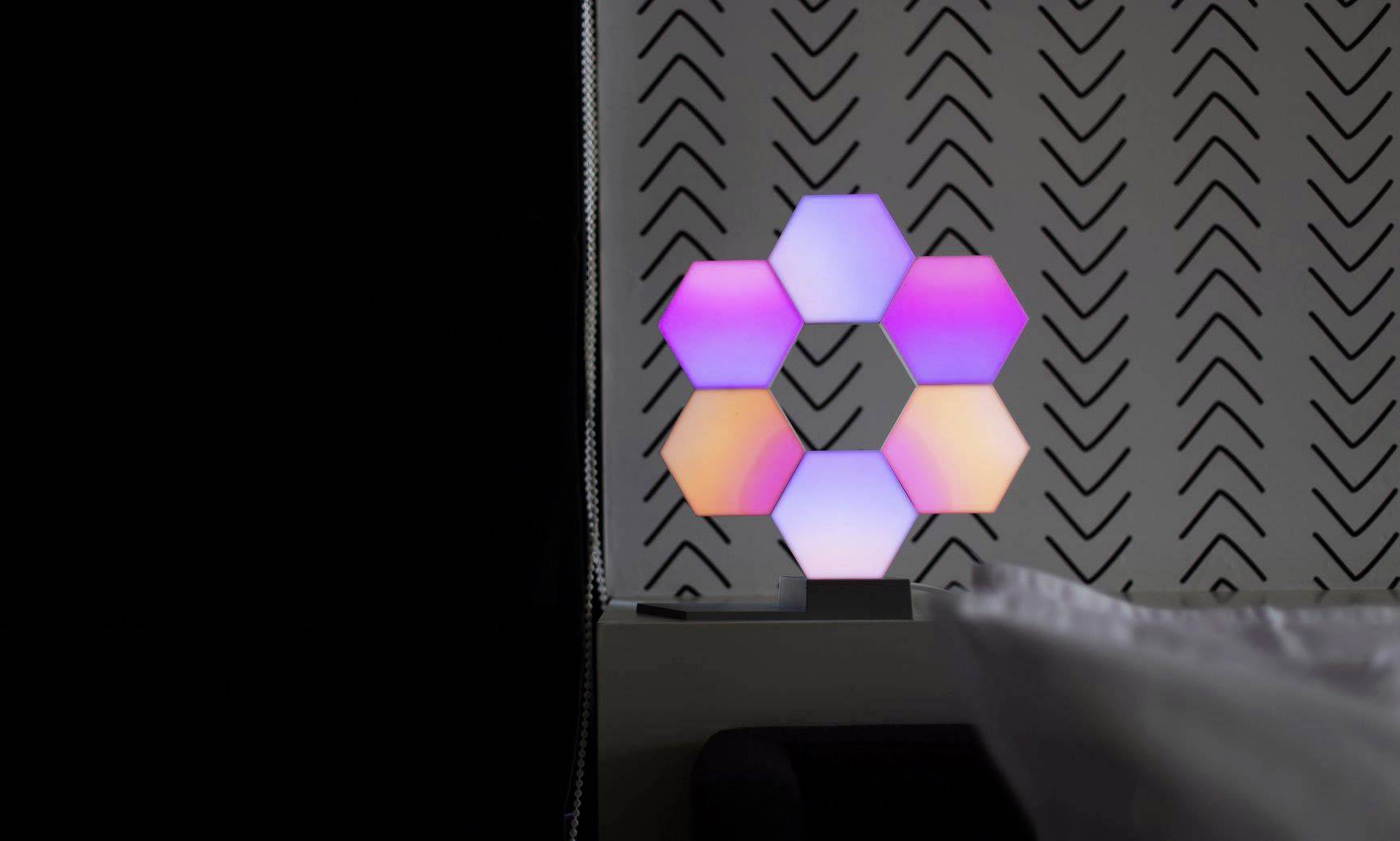 Sześciokątne lampy LED w pastelowych kolorach świecą na stole przed ścianą we wzory.
