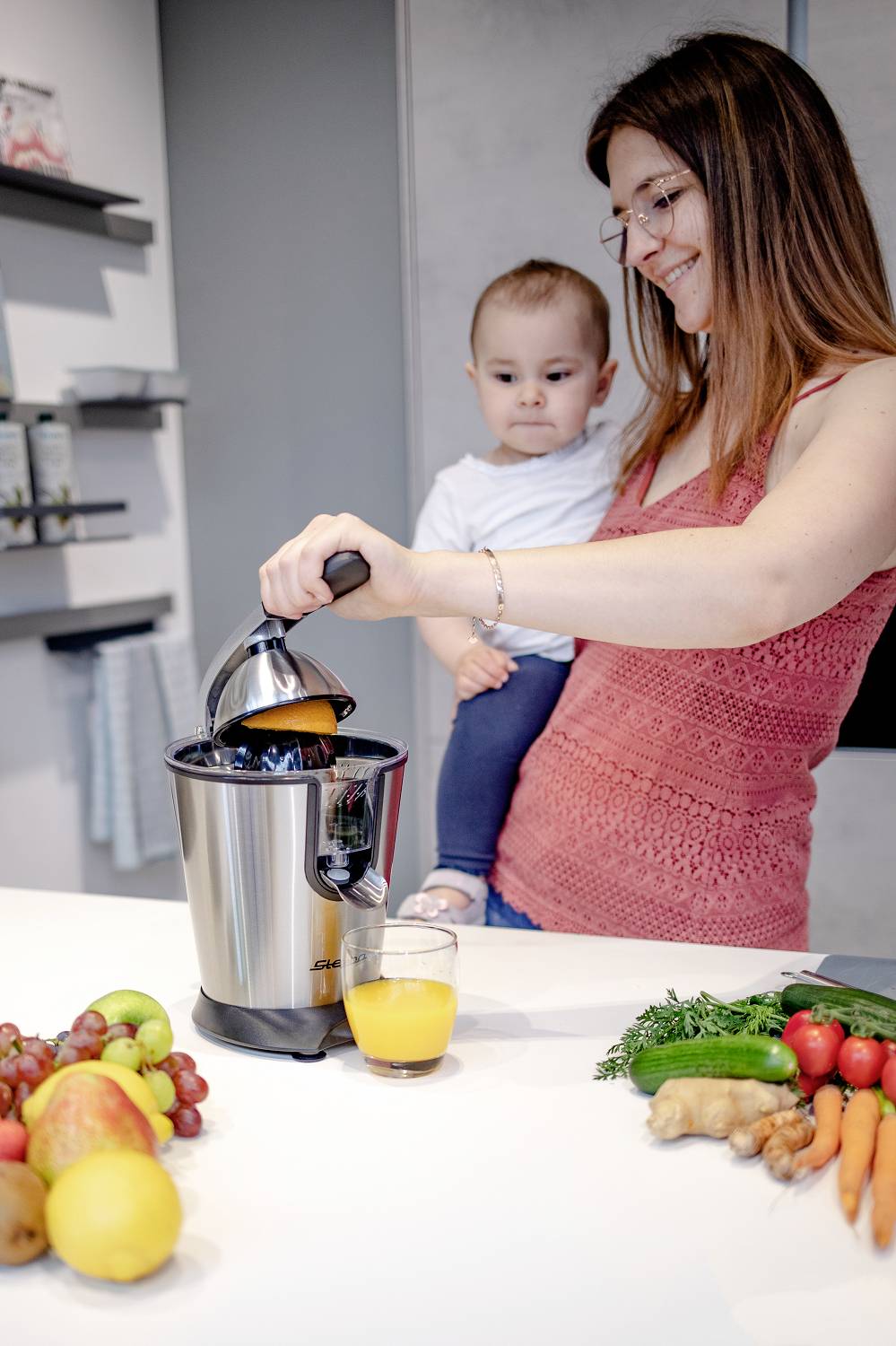 Kobieta trzyma niemowlę na ręku i wyciska pomarańcze sokowirówką. Na stole leżą owoce i warzywa.