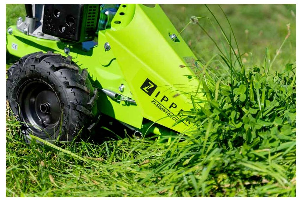 Zbliżenie kosiarki w kolorze zielonym, która kosi bujną trawę, ukazujące jej czarne koło i ostrza w ruchu. Nazwa marki 'Zipper' jest widoczna.