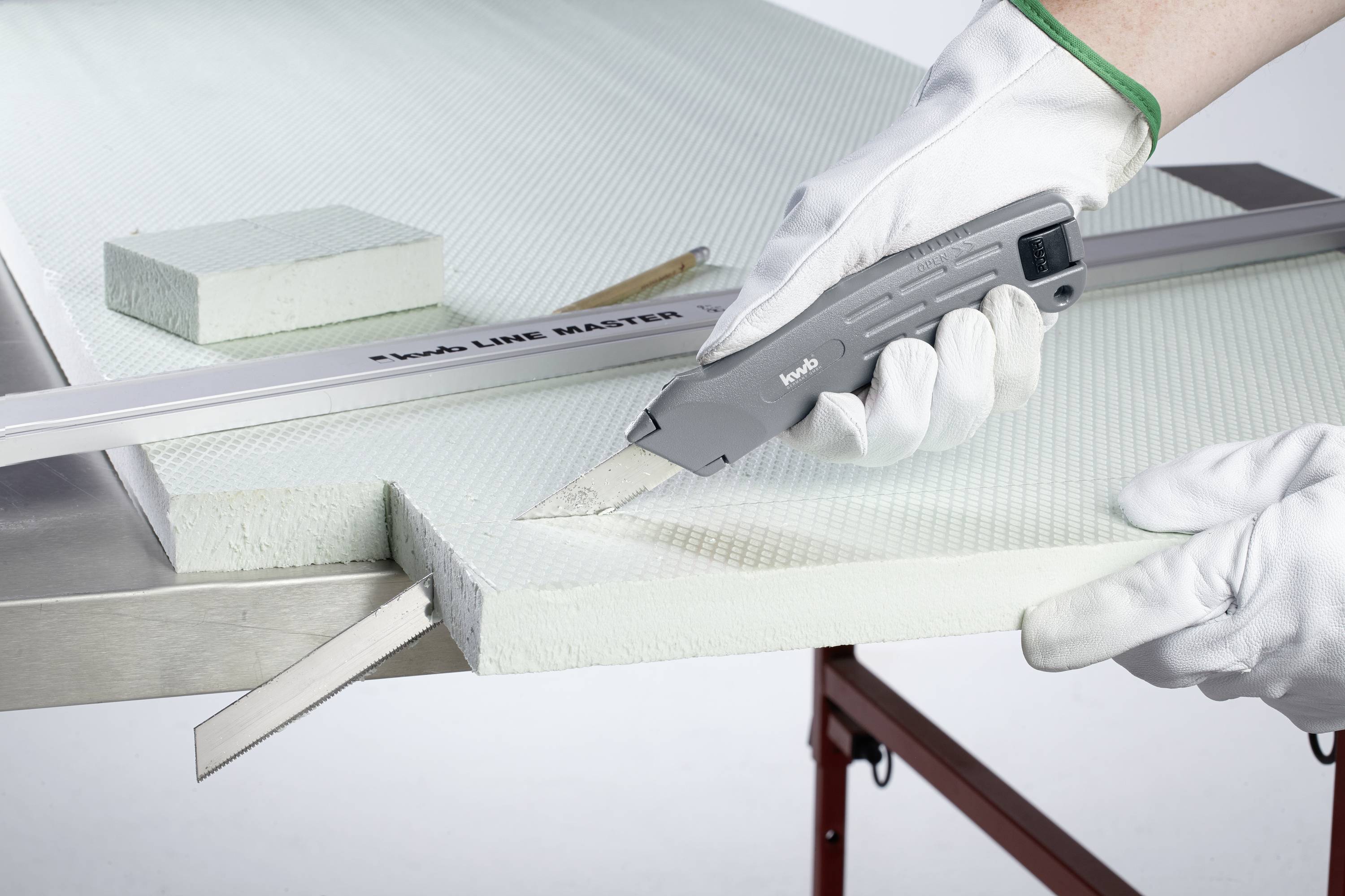 Osoba w białych rękawiczkach przecina nożem jasną płytę izolacyjną leżącą na stole.