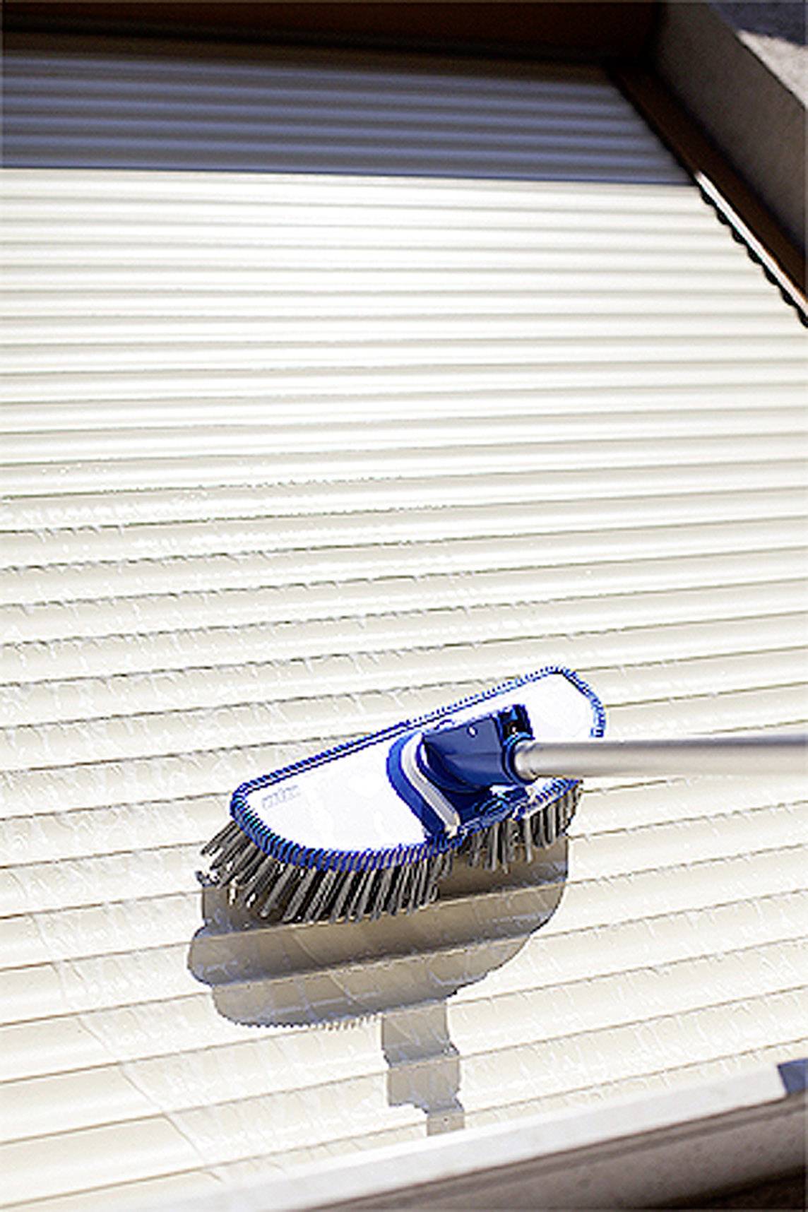 Okiennica jest czyszczona pędzlem. Pędzel posiada białe włosie i niebieską rączkę. Światło słoneczne rzuca cienie.