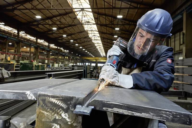 Pracownik w odzieży ochronnej używa elektronarzędzia do cięcia metalu w przemysłowym magazynie, otoczony stalowymi belkami i jasnym oświetleniem.