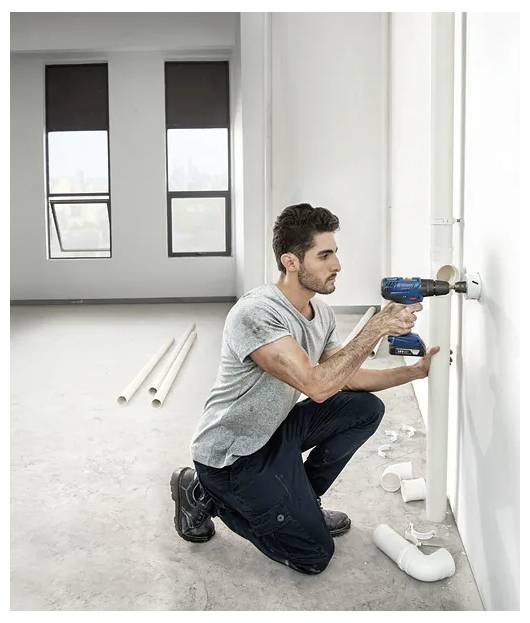 Mężczyzna klęczy na podłodze, używając wiertarki elektrycznej do naprawy rury w jasnym, nieukończonym pomieszczeniu. W pobliżu rozrzucone są plastikowe rury i złączki.
