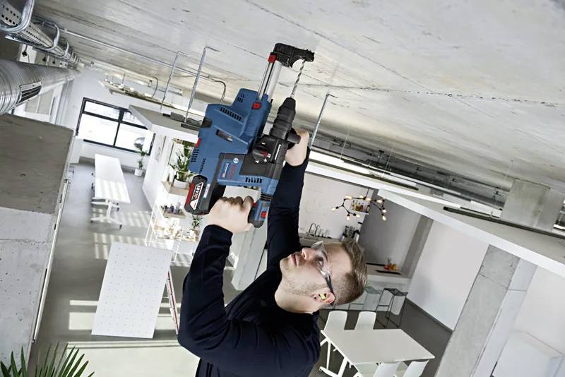 Osoba w okularach ochronnych używa wiertarki elektrycznej na betonowym suficie w nowoczesnym, industrialnym pomieszczeniu z dużymi oknami i stołami.