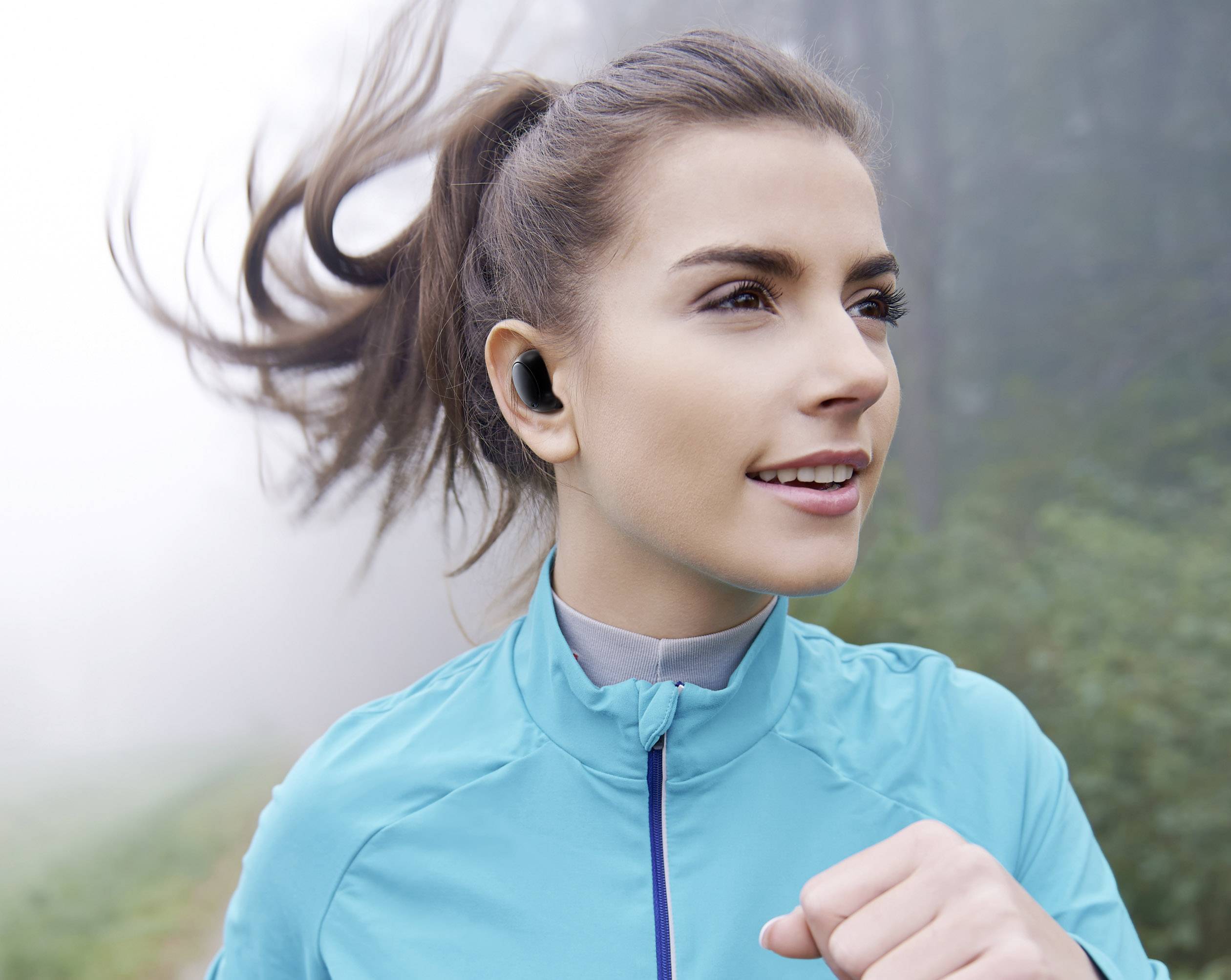 Kobieta biegnie w mglistą pogodę, ubrana w niebieską kurtkę i bezprzewodowe słuchawki douszne, patrząc przed siebie z koncentracją.