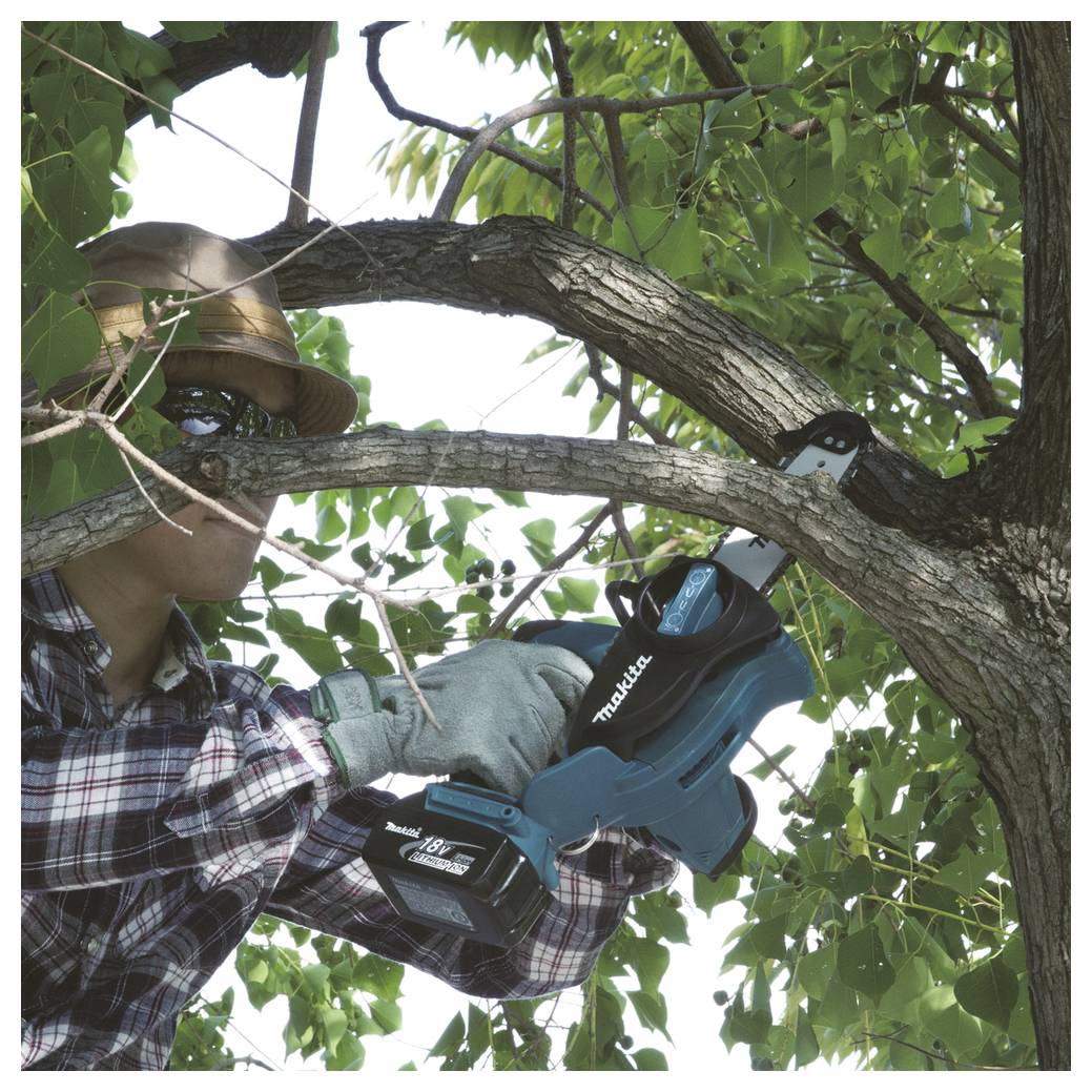 Osoba w kapeluszu i koszuli w kratę używa niebieskiej bezprzewodowej piły Makita do cięcia gałęzi drzewa w bujnym otoczeniu roślinnym.