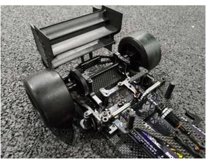 Podwozie samochodu RC bez nadwozia na asfalcie, eksponujące duże tylne koła i tylny spoiler, podkreślające techniczne szczegóły i strukturę.