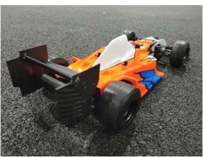 Mały, pomarańczowo-niebieski model samochodu wyścigowego stoi na nawierzchni asfaltowej, z charakterystycznym skrzydłem tylnym i czarnymi kołami.