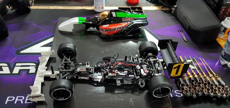 Zdalnie sterowany samochód w procesie budowy na stole warsztatowym, otoczony narzędziami i częściami samochodowymi. Główna bryła pojazdu spoczywa pośrodku.