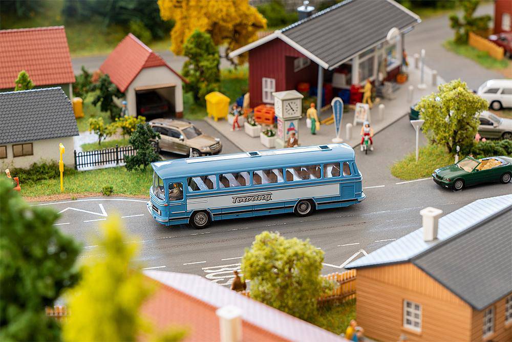 Miniaturowy model przedstawia niebieski autobus jadący po drodze w otoczeniu wiejskiej zabudowy, z małymi domkami i drzewami w tle.