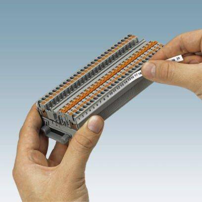 Osoba trzyma szarą listwę zaciskową z pomarańczowymi dźwigniami, używaną do elektrycznego łączenia przewodów.