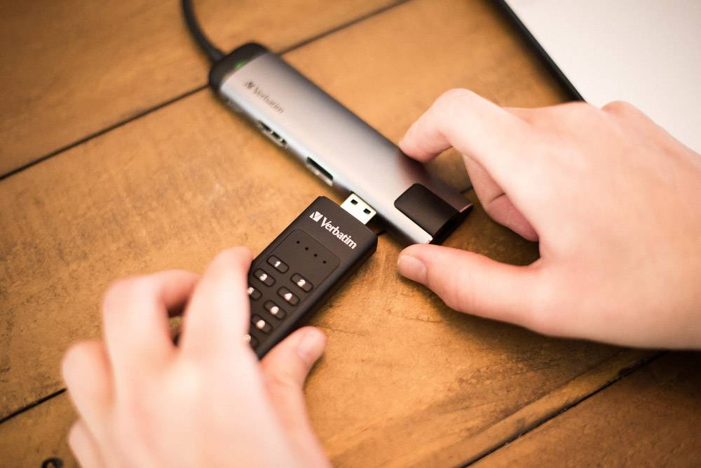 Dwie ręce podłączają napęd USB do adaptera. Na stole leży pendrive marki Verbatim.