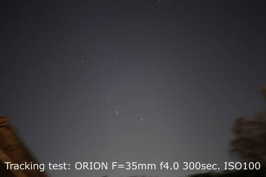 Nocne niebo z gwiazdami. Nieostre korony drzew po lewej stronie u dołu. Tekst u dołu: 'Test śledzenia: ORION F=35mm f4.0 300s. ISO100'.