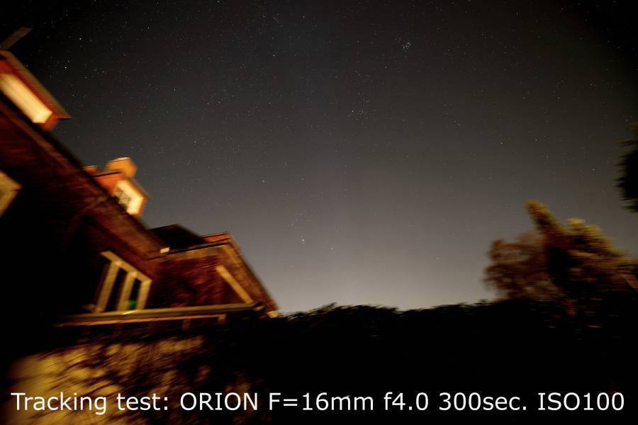 Nocne niebo nad budynkiem. Rozmyty firmament gwiaździsty na zdjęciu z tekstem 'Test śledzenia: ORION F=16mm f4.0 300sek. ISO100'.